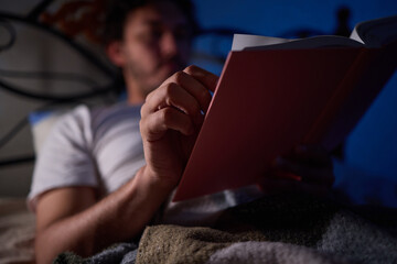 A man is peacefully reading a book in bed in a dimly lit bedroom, covered with cozy blankets