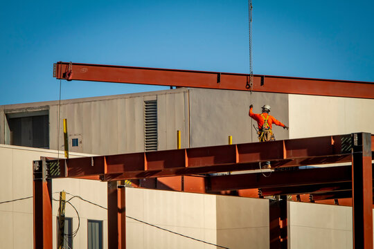 A wide shot of a construction worker directing placement of a large steel beam that is atttached to a crane