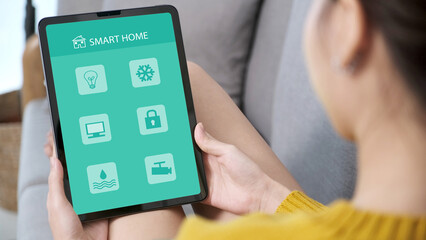 Hand using digital tablet with smart home control icon on screen while sitting on sofa at home, smart home control concept
