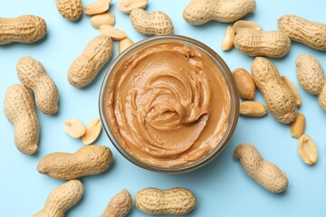Tasty peanut butter in bowl and groundnuts on light blue background, flat lay
