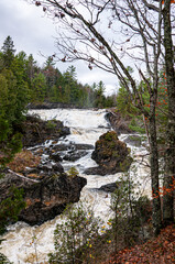 Nestled in the wilds of Quebec, the Chutes du Moulin cascade powerfully through rocky terrain, framed by early-autumn foliage and evergreens, under a cloudy sky.