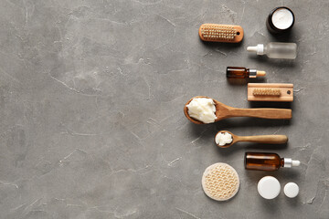 Spoons of shea butter with bottles of cosmetic oil and massage brushes on grey background