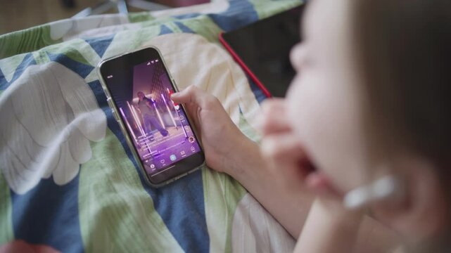 Caucasian girl surfs Internet spending leisure time in bedroom. Teenager lies on bed, watches video content in social network using mobile phone. Dancing young girl displayed on phone screen. Close up