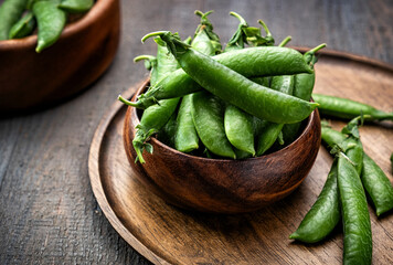 A handful of fresh green pea pods in a walnut wooden bowl, on a wooden round tray, on a gray textured wooden table. Rustic. Low key.