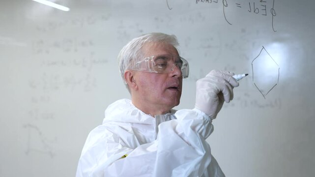 An elderly Caucasian male chemist in a protective suit writes on glass. 