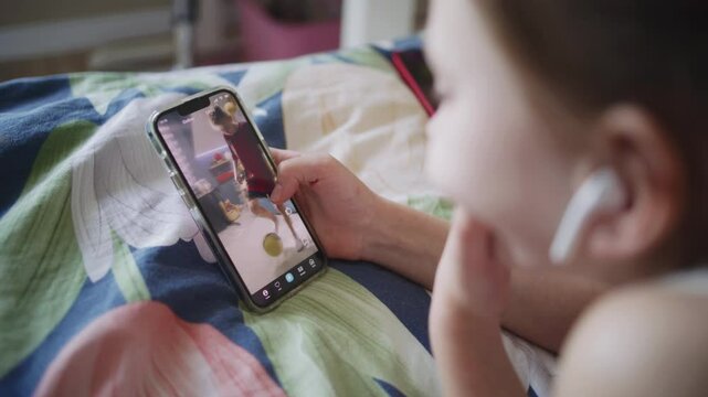 Close up shot of young girl lying on bed at home, surfing Internet and scrolling social networks using her mobile phone. Caucasian teenager spends leisure time in comfort bedroom. Lifestyle concept.