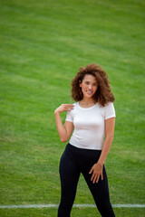 A beautiful curly girl posing in a white T-shirt and black leggings. Against the background of the stadium. Suitable for mockup