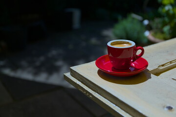 A full red espresso cup on an old white wooden table with green plants in the bokeh background.