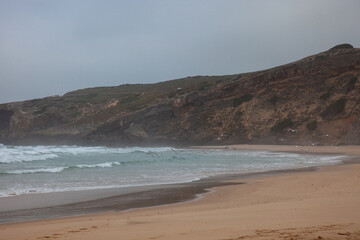 Fototapeta premium Monte Clérigo beach in Aljezur, Portugal, on a foggy morning. Seagulls take flight from the sandy shore, creating a serene and mystical coastal scene.