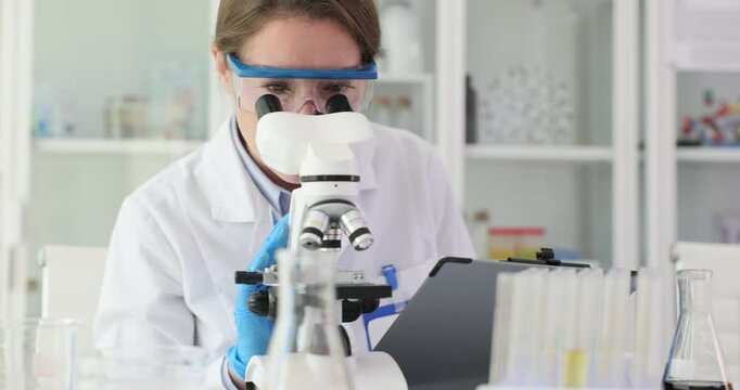 Female lab worker looks in microscope making notes. Medical researcher examines specimens recording detailed observations in notebook