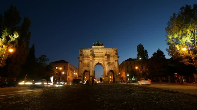 A large archway with a statue of a man on top of it. The archway is in front of a building with a clock tower. The sky is a beautiful blue color