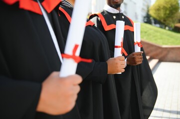 Group of happy smiling multicultural people in graduation gowns and caps with diplomas in hands graduate university outdoors in campus looking at camera. Graduation from college, university concept.