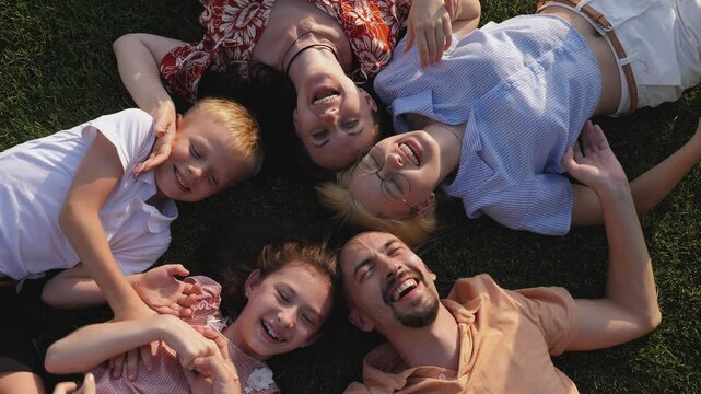 A big happy family lies in a circle on a green lawn in the park in summer and laughs. Brother tickles his sister. View from above.