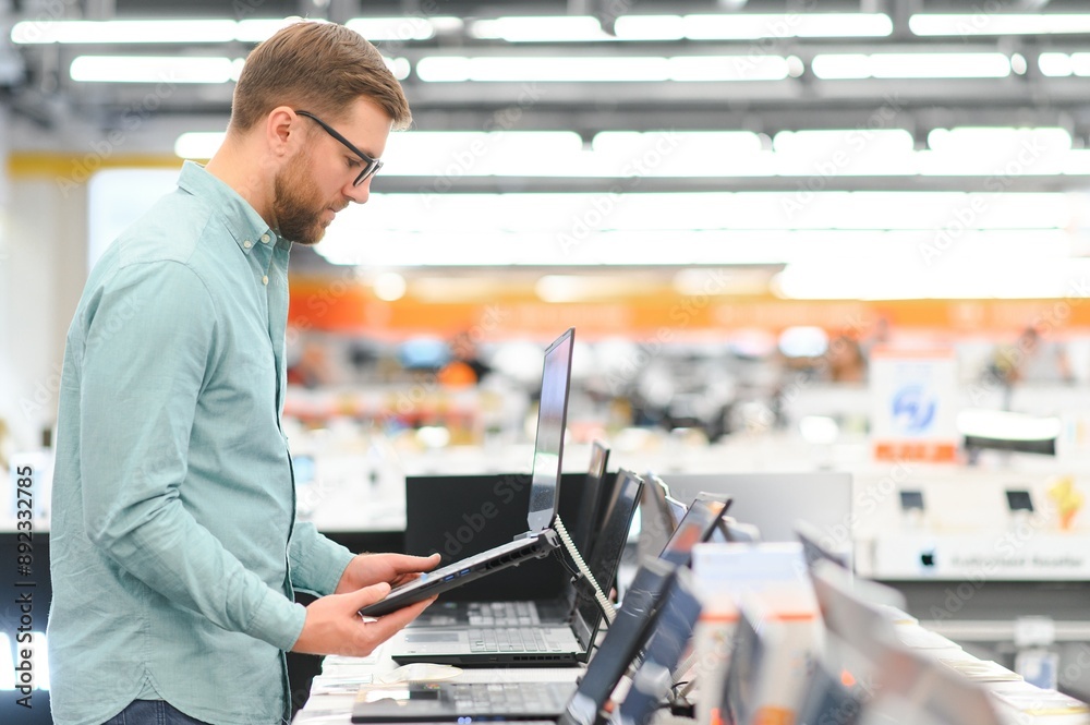 Wall mural a young man stands behind his laptop at the electronics store. a young man chooses a laptop in the s