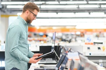 A young man stands behind his laptop at the electronics store. A young man chooses a laptop in the store