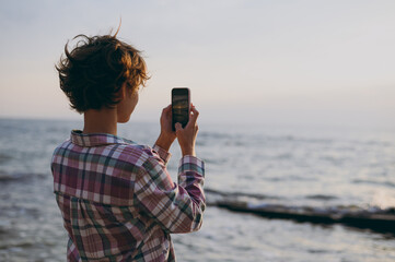 Back rear view young woman she wear shirt casual clothes taking photo of sunset sky use mobile cell phone rest on sea ocean sand shore beach outdoor seaside in summer day free time. Lifestyle concept.
