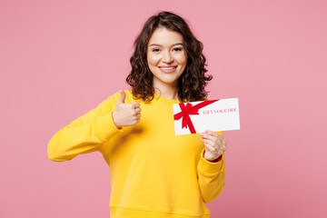Young woman she wearing yellow sweatshirt casual clothes hold gift certificate coupon voucher card for store show thumb up isolated on pastel light pink background studio portrait. Lifestyle concept.