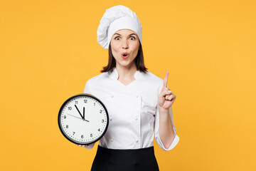 Young smart happy chef cook baker woman wear white shirt black apron uniform toque chefs hat hold in hand clock point index finger up isolated on plain yellow background studio. Cooking food concept.