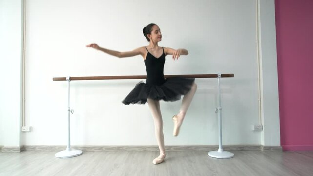 Young beautiful fourteen year old girl in black ballet tutu practicing ballet dancing practicing dancing as a black swan using bar pole, white wall in studio