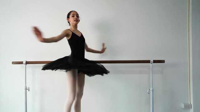 Young beautiful fourteen year old girl in black ballet tutu practicing ballet dancing practicing dancing as a black swan using bar pole, white wall in studio