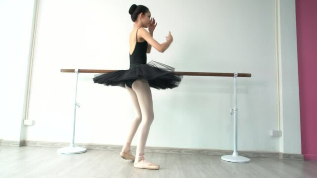 Young beautiful fourteen year old girl in black ballet tutu practicing ballet dancing practicing dancing as a black swan using bar pole, white wall in studio