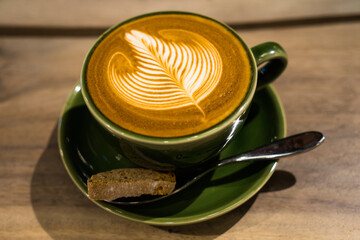 Hot latte with amazing latte art in bottle-green ceramic cup and saucer on wooden table with spoon and cupcake