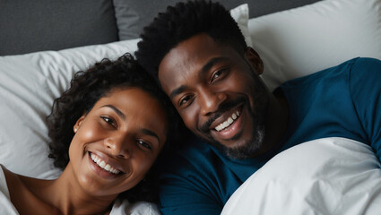 Fototapeta premium Happy Black couple lies in bed together, smiling at camera. They are both dressed in casual attire and appear to be relaxed and enjoying each other's company