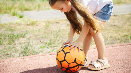 Child girl playing football at stadium. Little caucasian child ready to kick soccer ball at green field outdoors. Summer activities for children and future athlete concept.