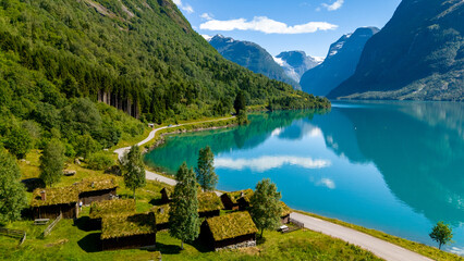 Traditional Norwegian cabins with grass roofs nestled by a serene fjord, surrounded by lush green...