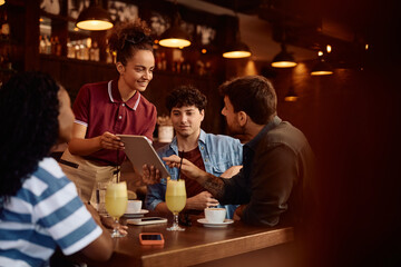 Happy waitress showing menu on touchpad to group of guests in  bar.