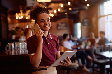 Happy waitress talking on cell phone while using digital tablet in  pub.