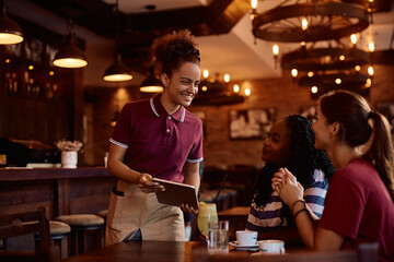 Happy waitress using touchpad while serving female guests in  pub.