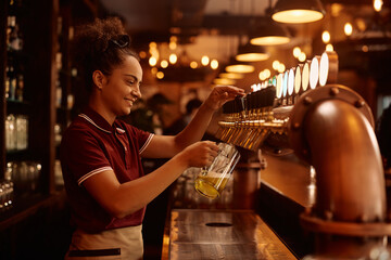 Young female barista pouring draft beer from beer tap in pub.