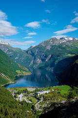 A charming Norwegian village set amid majestic mountains and a glistening fjord. Greenery blankets the land, buildings dot the shore, under a clear blue sky, Geiranger Fjord Norway