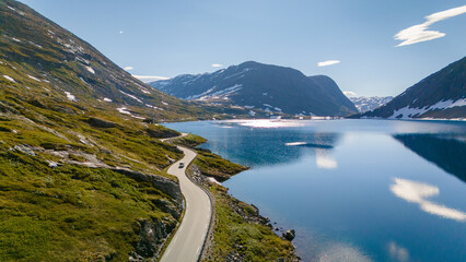 A single car winds its way through the majestic Norwegian landscape, a stunning lake reflecting the sky and snow-capped peaks. Langvatnet, Geiranger, Norway