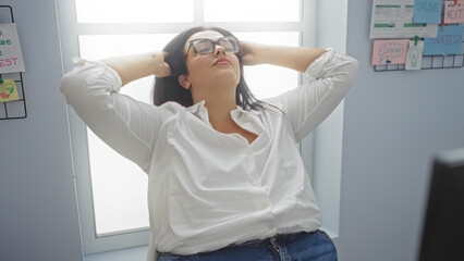 Woman relaxing in an office setting, demonstrating a sense of relief while leaning back in her chair with her hands behind her head.