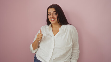 Young hispanic woman giving thumbs up in front of isolated pink background, smiling confidently while wearing white blouse and jeans, representing body positivity