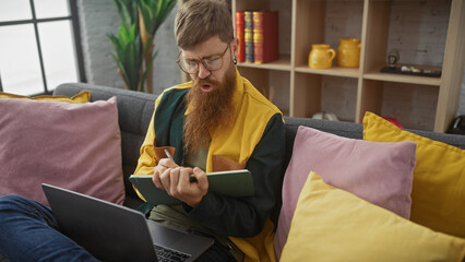 Redhead bearded man with glasses writing in notebook on sofa, focused, using laptop, indoors, casual