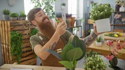 Obraz premium Smiling bearded man taking a selfie with a plant in a lush indoor flower shop.