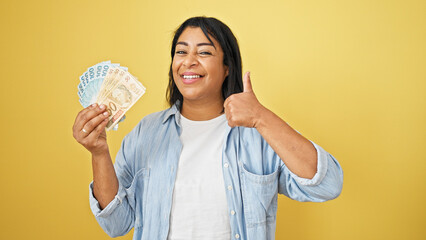 Hispanic woman smiles holding brazilian real, thumbs up on yellow background