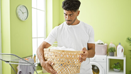 A handsome hispanic man holds a laundry basket in a bright green home laundry room with modern...
