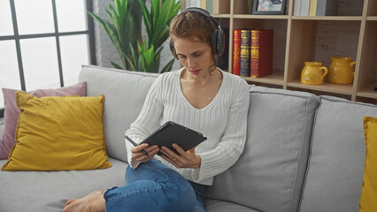 Obraz premium A young woman wearing headphones uses a tablet while sitting on a sofa in a cozy living room.