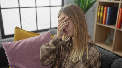 A young blonde woman indoors covers her eyes with her hand, showing distress against a cozy home background.