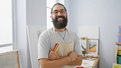 Fototapeta premium Hispanic artist with beard and apron smiling confidently indoors surrounded by paintings in a studio atmosphere.