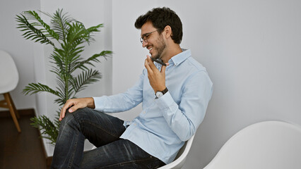 Handsome hispanic man laughing while listening to a voice message in a modern waiting room.