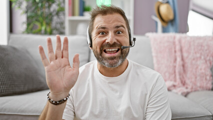 Hispanic mature man with grey hair in a casual white t-shirt waves cheerfully during a video call from a cozy living room.