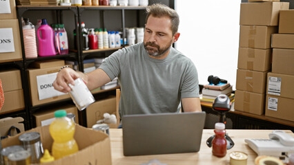 Hispanic man organizing cans in a donation center warehouse while working on a laptop.