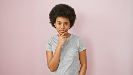 Pensive african american woman with curly hair posing against a pink isolated background in a casual gray shirt.