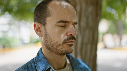 Fototapeta premium Close-up of a contemplative bearded bald man with eyes closed in a serene park setting surrounded by greenery.