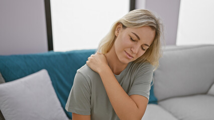 A beautiful blonde woman experiences discomfort in a modern living room, touching her neck gently.